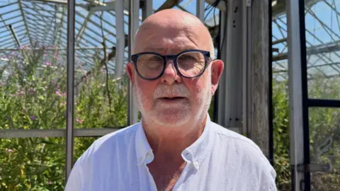 Kevin Herve pictured in front of a derelict glass house. He is wearign a white shirt and glasses. 
