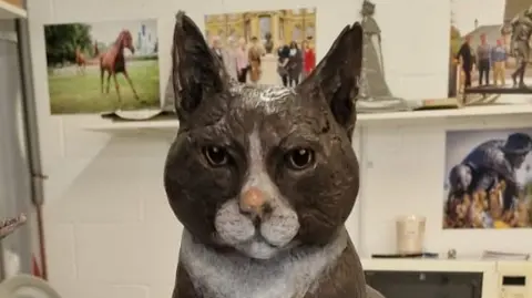 A life-size statue of a black and white cat sitting on a metal plinth. The statue is in an art studio and there are photos of other statues on the wall behind it.