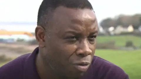 Leroy Rosenior. He has short dark hair. He is looking to the side of the camera. 