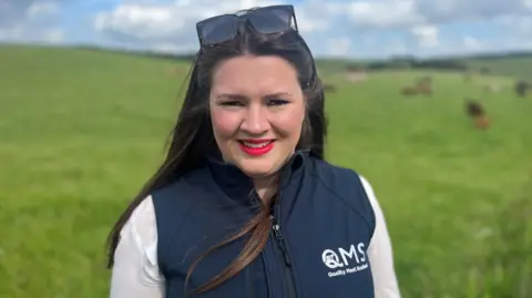 Sarah Millar, wearing a white shirt and blue gilet marked with the QMS logo, looks to the camera at the side of a field.  She has dark brown hair, red lipstick and has sunglasses on her head.