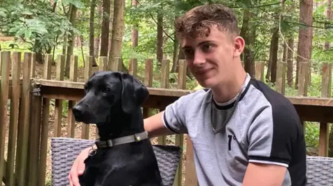 Norfolk Police Image of Cory Chilvers sitting with his arms around a black Labrador dog. He has light brown hair and is wearing a grey T-shirt. He has a silver chain around his neck.