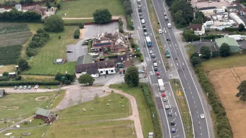 Rees Keene - Over Farm A bird's eye view of traffic is pictured on a carriageway, with surrounding fields and houses.
