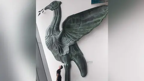 Arts Groupie A young boy stands underneath and looks up to a huge Liver Bird sculpture in a building. 