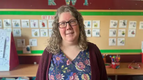 Mum Ruth stands in a classroom wearing a floral print top and burgundy cardigan. She has shoulder-length curly hair and glasses.