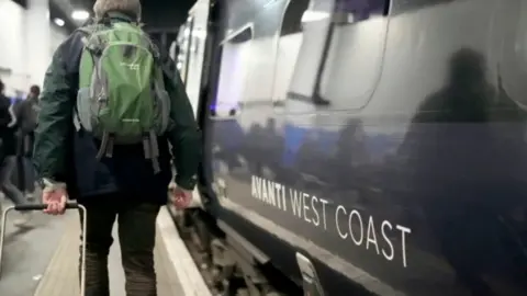 A man with a green backpack on is pulling a case on a platform alongside an Avanti West Coast train
