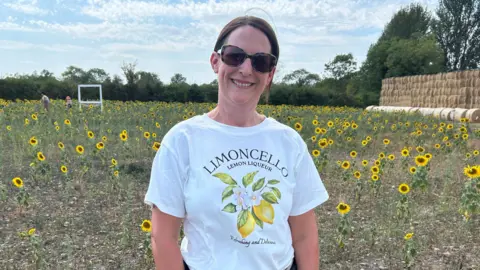 BBC A smiling woman with brown hair, tied back, wearing sunglasses and a white t shirt with lemons on it. She is stood in a field of sunflowers which are no taller than waist height.