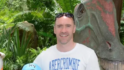 Paul Doyle wearing a white t-shirt, smiling with sunglasses on his head.