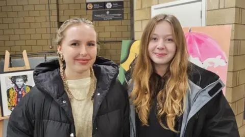 The image shows Natasha and Neve Bowater standing side by side indoors, likely at an event or exhibition. Both are wearing coats, and one is holding a drink and some papers. Behind them are display boards or easels featuring artwork, including a colourful portrait and a painting of a pink umbrella. The setting appears to be a large hall with brick walls and signage in the background.