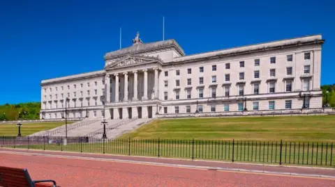 Getty Images Stormont is a large sandstone building with windows and a large set of steps leading up to it. There is a black metal fence around the gardens and a bench sitting on the footpath.