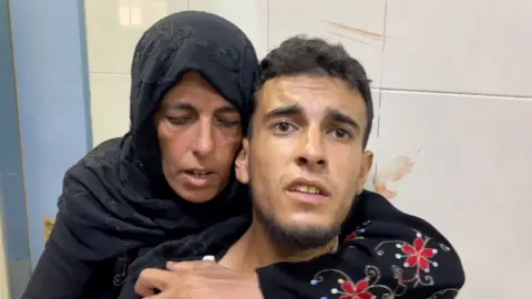 A woman wearing a black headcovering leans over the shoulder of a younger man with dark brown hair. They both look distressed and there looks to be smears of blood on the walls behind them.