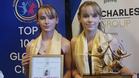 A pair of teenage girls who are identical set of twins, both blonde and wearing gold scarves over their necks, holding up a gold award and a framed certificate.
