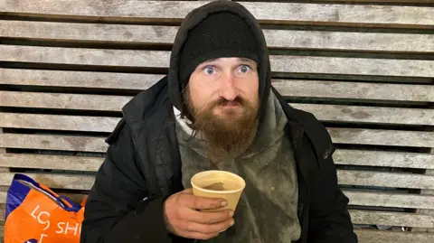 Rough sleeper Lee, who the street pastor team met at Southwater.  Lee is sitting on the ground holding a cup of tea and is leaning against a fence.  He's wearing a thick coat and fleece along with a hat.  He has a plastic bag of belongings sat beside him.