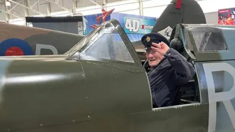 Submitted An elderly man is sitting in the cockpit of a Spitfire, wearing a peaked cap and saluting while he looks at the camera