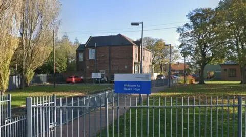 Rose Lodge in Hebburn. The ward is a converted detached house. A paved path leads to the front doors of the property. Green lawn lines either side of the path. The building is located behind a metal fence and a welcome sign.