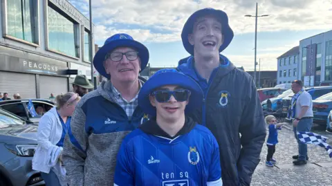 Dungannon Swifts fans Eddie, Mathew and Phillip. Eddie is wearing a blue hat, glasses, check shirt and Swifts jacket. Matthew is wearing a blue hat, sunglasses and blue swifts jersey. Philip is wearing a blue hat and black jacket.