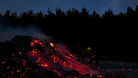 It is night and a firefighter is cast in a red glow from a mound of burning muck that looks almost like lava from a volcano. The firefighter is using a hose to spray the mound and is wearing a yellow helmet. The sky is a dark night blue and the trees around the edges of the farm and are cast as silhouettes. 
