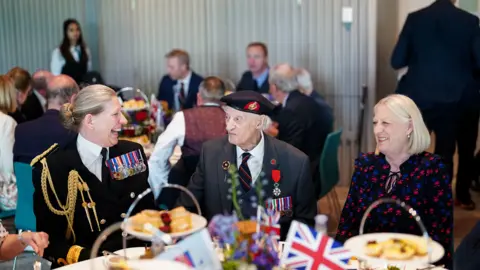 Two women and a man sit at a table with flags and cakes as a tea party is held at the National Memorial Arboretum during VE Day.