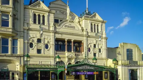 Manx Scenes An ornate, Victorian-era theatre with a cream frontage. "Gaiety Theatre and opera house" is written in letters on the third floor, along with a greek statue of a woman holding a touch aloft. On the ground level there is a steel and glass canopy above the entrance.