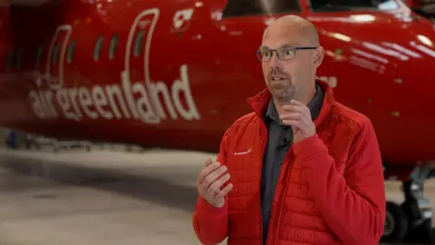 El presidente de Air Greenland, Jakob Netter Sorensen, siendo entrevistado delante del avión y vestido con una chaqueta roja.