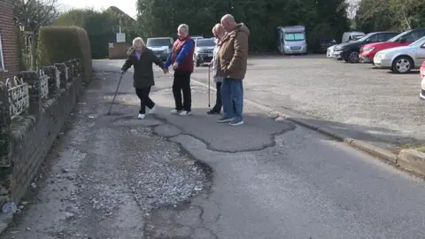 Four elderly people are standing in the road riddled with potholes. There are two women, both holding walking sticks, with their husbands holding their hands. They are all wearing warm winter coats and one man is pointing at the potholes in the road.