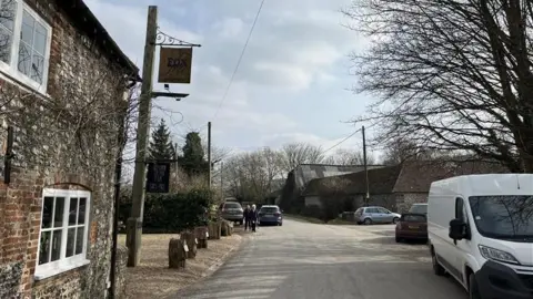 George Carden/BBC The Fox Goes Free pub is a flint and brick walled pub with small windows and low door frames. It is next to the main narrow lane through the village which has cars parked on it. And there is a barn on the other side of the road