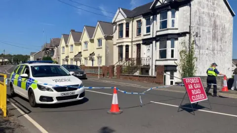 A police car is seen along a road, with police tape and a red "Road Closed" sign