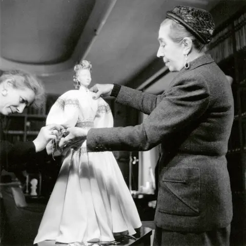 Vitra Design Museum Archive A monochrome image of two fashion ladies fixing a pale dress onto a small doll mannequin. They are dressed in 1940s-style wool suits.