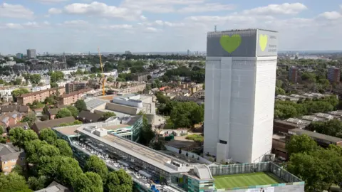 PA Media Drone view showing Grenfell Tower covered entirely in white with a green heart at the top. It rises above buildings, a sports pitch and trees with the London skyline behind.