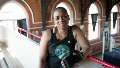 Antonia Amja Lee leans on a red railing inside a historic boxing venue, wearing a black Fight Forward vest and a black hand wrap.