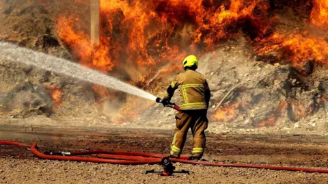A large outdoor fire burning across what appears to be a heap of hay. Thick orange and red flames stretch across much of the scene, while dense grey smoke rises high above the fire and partially obscures the background.

In the foreground, a firefighter stands facing the blaze and is operating a high-pressure hose. The firefighter is wearing protective firefighting gear, including a brown fire-resistant suit with reflective yellow stripes and a bright yellow helmet. A powerful stream of water is being directed from left to right into the heart of the fire.