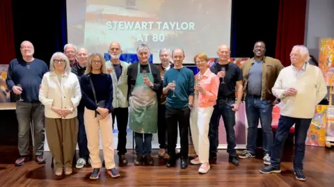 Thirteen people stand with a glass of fizz in their hand. Paintings can be seen behind them and a projection on a wall behind them says "Stewart Taylor at 80".