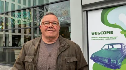 Gary Cross standing outside the Transport Museum in Coventry. He has short grey, black hair and wears a dark green jacket over a grey top. He has red-rimmed glasses on.