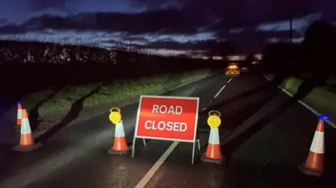 A straight section of country road pictured at night. In the foreground are cones and a sign stating, 'Road Closed'. Emergency vehicles are in the distance.