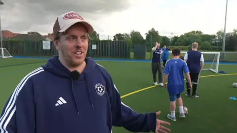 A person in a cap and navy sports hoodie stands on an outdoor football pitch, speaking while gesturing with one hand. In the background, several people in sportswear walk and handle a football near a small goal on the artificial turf. The scene takes place under a cloudy sky, with fenced surroundings and trees visible around the field.