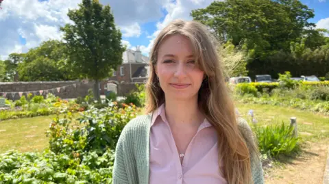 Annabel pictured in a public garden. She has long blonde hair and is wearing a green cardigan and pink shirt. She 