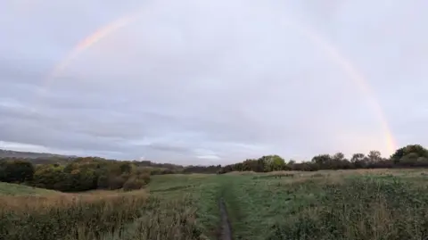 BBC A track leading into Crawcrook Quarry. The track leads into a large field filled with grass and shrubs. Woodland can be seen in the background. A rainbow arcs across the sky.