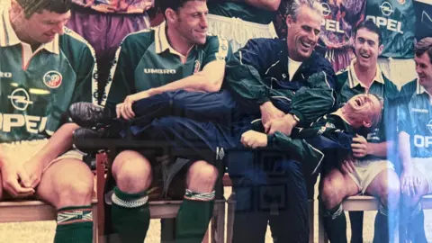 O'Leary family A photo of the 1990s Republic of Ireland team - four players and manager Mick McCarthy are sitting down for a team photo and Charlie O'Leary lies across them as McCarthy tickles him and he laughs. The players are wearing dark green shirts while McCarthy and O'Leary are in dark blue tracksuits with green arms.