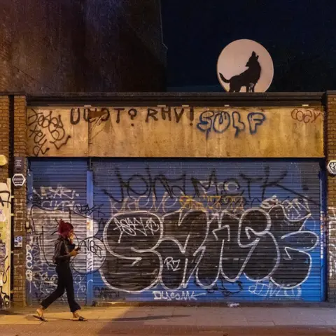 Banksy/Instagram Night-time image of black silhouetted wolf painted on satellite dish so it looks like a moon above a shop with blue shutters in Peckham Rye