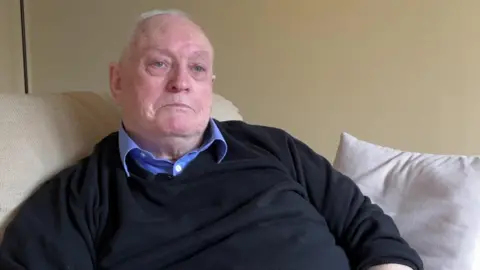 A man sat on a sofa wearing a black jumper and blue collared shirt.