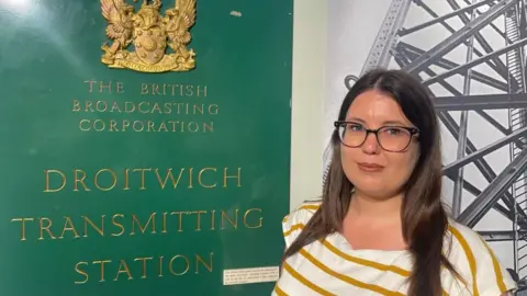 A woman stands to the right of the picture - she has long brown hair and glasses, wearing a mustard yellow striped t-shirt. She is standing in front of a black and white photo of a transmitter mast and to the left is a green and gold sign which says The British Broadcasting Corporation, Droitwich Transmitting Station.