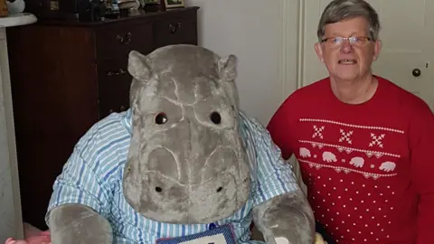 Philip Angier is a man in his 70s wearing a red Christmas jumper with brownish hair and glasses. He is sitting next to a huge hippo soft toy which is almost as big as he is 