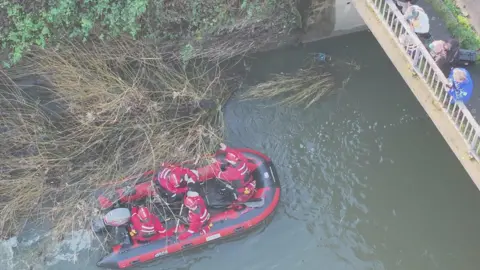 HARTSAR A drone image showing the red dinghy, with the four firefighters on it in red, getting the dog onto the boat. You can see some people to the right standing on a footbridge watching on.