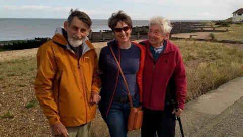 Colin, Christine and Beryl Bell are standing in front of a beach. Colin - an older, bearded man on the left of the three is smiling. He's wearing an orange raincoat. Christine, in the middle of the three, is wearing a blue coat and sunglasses. She has short brown hair. On the right, Beryl is an older woman in a red fleece coat. She is smiling at Christine and holding onto a walking stick.