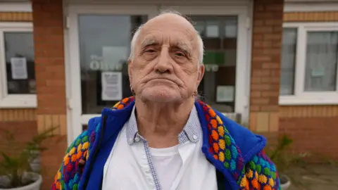 Qays Najm/BBC John Manning stands in front of The Olive cafe, looking expressionless at the camera. He is bald with some grey hair on the side of his head. He wears a white shirt with a colourful jacket over the top. 