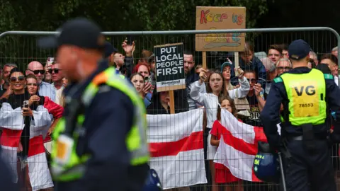 Protesters holding up England flags and placards behind a metal fence. Two police officers are watching on.