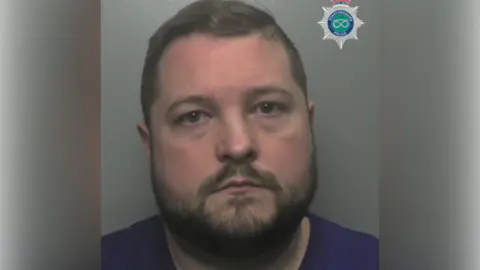 A police mugshot of a man with short dark hair, a beard and a blue shirt.
