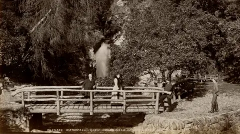 Manx National Heritage An early 20th century black and white photograph shows a footbridge made of logs across a river, with people walking over it. There are trees all around and a waterfall in the background.