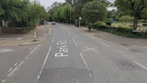 A view of Park Road taken from Google street view. It shows a road with trees on either side. There is one car in the distance approaching the camera.  