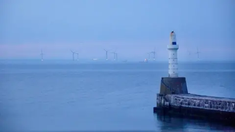 Getty Images A white lighthouse, with wind turbines and offshore supply vessels in the distance in the sea off Aberdeen, under a blue/pink sky.