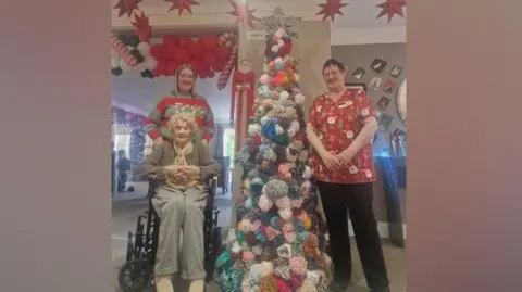 Grampian Court Care Home Lisa Stephenson, the home’s Wellbeing Co-ordinator is smiling next to the tree. She is wearing a red and green stripy Christmas jumper and has shoulder-length blonde hair. Resident Hylda Carr is in a wheelchair in front of her. She has short curly white hair and is wearing grey jumper and trousers and a gold scarf. On the other side of the tree is housekeeper Karen who supplied all the wool for the pompoms. She is wearing a red Santa T-shirt and black trousers. She has short red hair.